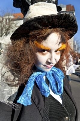 Eine Person in einer Hutmacher-Verkleidung mit schwarzem Hut, blauer Schürze und Gesichtsbemalung, die im Freien in einer Stadt steht.