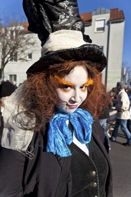 Eine Frau mit roten Haaren und orangefarbenem Augen-Make-up ist als Mad Hatter gekleidet, trägt einen schwarzen Hut mit weißer Schleife, ein blaues Halstuch und einen schwarzen Mantel. Sie steht auf einer überfüllten Straße.