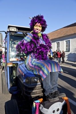 Eine Person in einem bunten Kostüm mit lila Federn sitzt auf einem blauen Traktor, möglicherweise an einer Parade teilnehmend. Dahinter steht ein Gebäude mit mehreren Fenstern und draußen stehenden Personen.