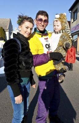 Eine Familie aus drei Personen, in Kostümen gekleidet, steht auf einer Straße mit einem Gebäude im Hintergrund. Der Mann trägt ein gelbes Shirt mit einer Nummer, die Frau einen Pelzmantel und das Kind ein Leoparden-Kostüm.