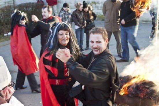 Eine Gruppe von Menschen in Halloween-Kostümen tanzt und lächelt im Freien. Ein Mann und eine Frau in der Mitte halten Händchen. Im Hintergrund brennt ein Feuer auf dem Kopf einer Person.