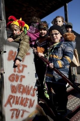 Eine Gruppe von Kindern in Winterkleidung und Mützen, einige mit Gesichtsbemalung, stehen auf einer Plattform mit Graffiti, das 'Bist gut wenns rinnen tut' besagt. Sie halten sich an einem Geländer fest und scheinen auf einem Umzugswagen zu stehen.