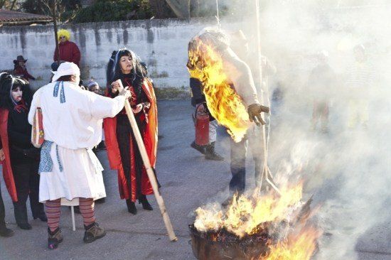 Eine Gruppe von Menschen in Kostümen versammelt sich um ein Feuer. Eine Person hält eine brennende Puppe, während eine andere Person einen Stock hält.