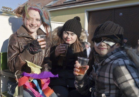 Drei junge Frauen in Winterkleidung und mit Gesichtsbemalung sitzen zusammen und halten Plastikbecher mit Getränken. Eine hat bunte Bänder auf der Schulter. Sie lächeln und scheinen einen festlichen Anlass zu genießen.