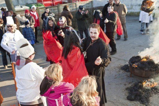 Eine Gruppe von Menschen in roten und schwarzen Kostümen, möglicherweise für eine Halloween-Veranstaltung, versammelt sich um ein Lagerfeuer. Ein Mann in einer Kochmütze steht in der Nähe.