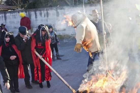 Eine Gruppe von Menschen in Kostümen versammelt sich um ein Lagerfeuer, wobei eine Person mit Maske und Handschuhen einen Stock hält.