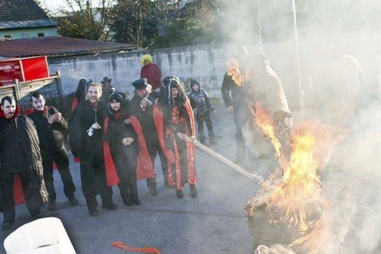 Eine Gruppe von Menschen in Kostümen steht um ein Lagerfeuer herum, eine Person hält einen Stock und pustet in das Feuer.