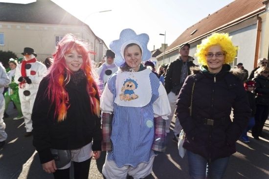 Drei Frauen in Kostümen, eine mit einem Babylätzchen, lächeln und posieren für ein Foto auf einer Straße. Dahinter laufen andere Menschen in Kostümen.