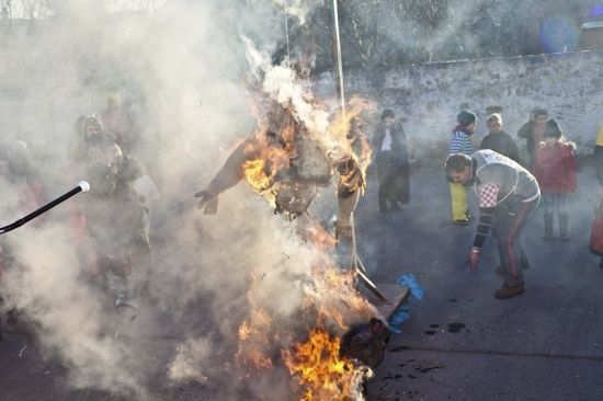 Eine große Puppe wird in einem Außenbereich verbrannt, während mehrere Menschen zusehen. Die Puppe ist in Flammen aufgegangen und Rauch steigt von ihr auf. Ein Mann steht in der Nähe, möglicherweise zur Überwachung des Verbrennens.