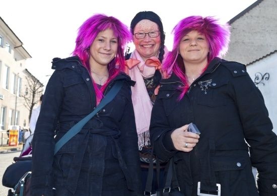 Drei Frauen mit rosa Haaren, lächelnd und stehend vor einem Gebäude. Die Frau links trägt eine schwarze Jacke und hat eine blaue Tasche. Die mittlere Frau trägt einen rosa Schal und eine Brille. Die Frau rechts hält ein Handy.