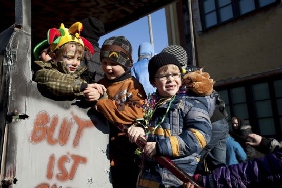 Drei Kinder in Festkostümen stehen auf einem Festwagen, halten eine Stange und lächeln. Der Wagen hat Graffiti darauf. Dahinter beobachtet eine Menge.