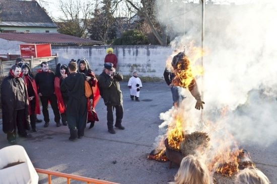 Eine Gruppe von Menschen in Kostümen versammelt sich um ein Lagerfeuer und beobachtet, wie eine Person auf einem Pfahl verbrannt wird.