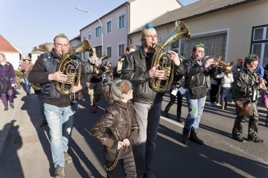 Eine Gruppe von Musikern, darunter zwei Männer, die Tuba spielen, gehen während eines Umzugs mit einem kleinen Kind in ihrer Mitte die Straße entlang.