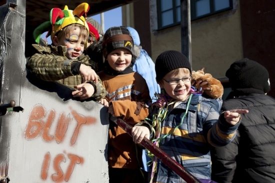 Drei Kinder in festlicher Kleidung, eines trägt einen Narrenhut, stehen vor einer mit Graffiti bedeckten Wand. Sie scheinen an einem Umzug teilzunehmen.