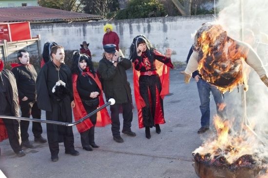 Eine Gruppe von Menschen in Kostümen, darunter eine Person in Flammen, stehen um ein Lagerfeuer herum.