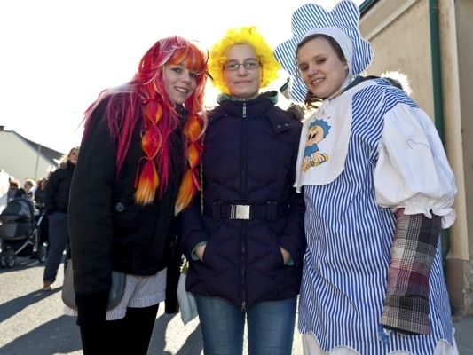 Drei Frauen in bunten Kostümen stehen zusammen draußen. Die Frau auf der linken Seite hat leuchtend pinkes und oranges Haar, die mittlere hat gelbe Haare und eine Brille und die Rechte trägt ein blau-weiß gestreiftes Kleid.