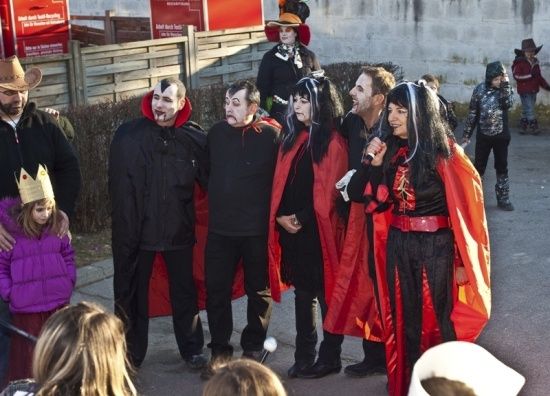 Eine Gruppe von Menschen in Vampirkostümen steht vor einer Wand. Einige halten Mikrofone und scheinen eine Aufführung zu veranstalten.