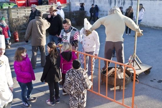 Eine Gruppe von Menschen, darunter Kinder, versammelt sich um ein Lagerfeuer mit einer großen Figur im Hintergrund. Einige tragen Kostüme, und ein Mann in einer Kochmütze hält einen Stock.