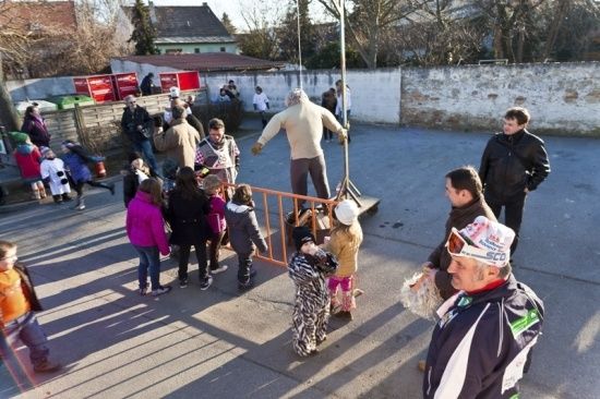 Eine Gruppe von Menschen, einschließlich Kindern, versammelt sich um einen Mann, der auf einer Plattform in einem Außenbereich steht. Der Mann ist in einem weißen Anzug und Handschuhen gekleidet. Die Kinder tragen verschiedene Winterkleidung. Der Bereich ist von einer Steinmauer und Bäumen umgeben.