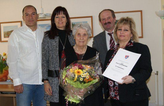 Fünf Personen stehen in einem Raum zusammen, wobei eine Frau einen Blumenstrauß und eine andere ein Zertifikat in der Hand hält. Die Frau in der Mitte ist älter.