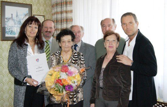 Eine Gruppe von Menschen steht in einem Raum zusammen, eine Frau hält einen Blumenstrauß und ein Zertifikat, während eine andere Frau ein Papier hält.