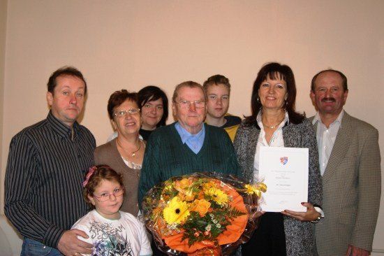 Eine Gruppe von Menschen steht lächelnd in einem Raum. Ein Mann in der Mitte hält einen Blumenstrauß. Eine Frau auf der rechten Seite hält ein Zertifikat.
