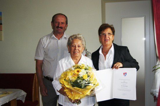 Drei Personen, zwei Frauen und ein Mann, posieren für ein Foto. Die Frau auf der linken Seite hält einen Blumenstrauß, während die Frau auf der rechten Seite ein Zertifikat hält. Sie alle lächeln.