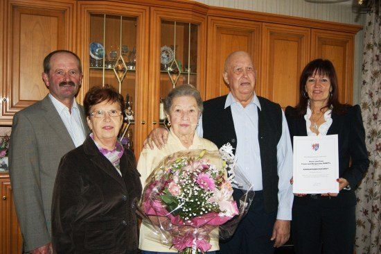 Eine Gruppe von Menschen, darunter eine ältere Frau mit Blumen, steht zusammen und lächelt für ein Foto. Dahinter befinden sich Holzschränke mit Glastüren.