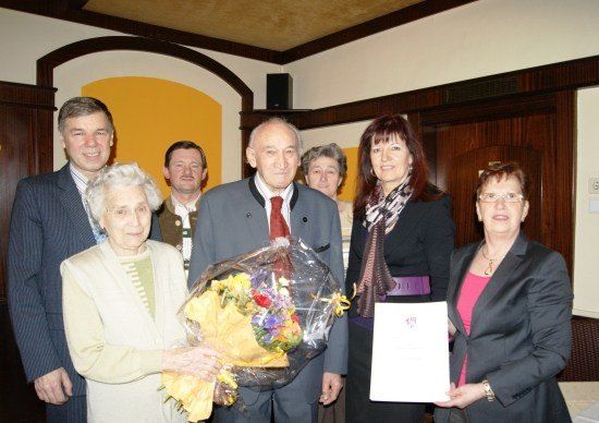 Eine Gruppe von Menschen steht zusammen, eine Frau hält einen Blumenstrauß und eine andere Frau hält ein Zertifikat. Sie alle lächeln und scheinen sich bei einer Veranstaltung zu befinden.