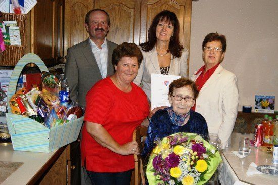 Fünf Personen stehen in einer Küche und lächeln für ein Foto. Eine Frau in einem roten Shirt hält einen Blumenstrauß und eine andere Frau mit Brille hält ein Zertifikat. Ein Korb mit Snacks steht auf der Theke.