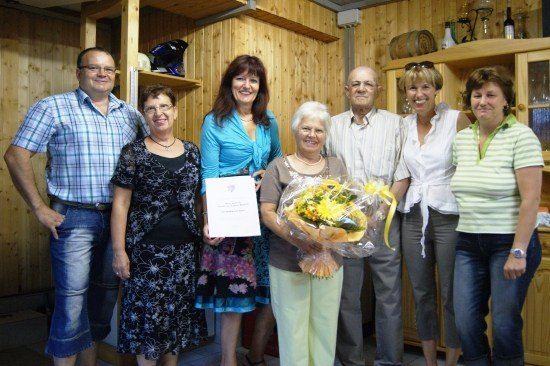 Eine Gruppe von Menschen posiert für ein Foto mit einer Frau, die einen Blumenstrauß und ein Zertifikat hält. Sie stehen in einem Raum mit Holzwänden und Regalen.