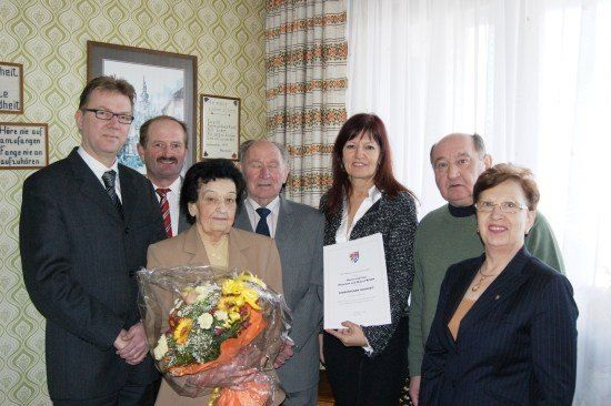 Eine Gruppe von Menschen steht in einem Raum, einer hält einen Blumenstrauß und ein anderer ein Zertifikat. Sie alle lächeln und scheinen für ein Foto zu posieren.