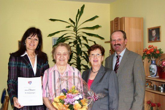 Vier Menschen stehen lächelnd in einem Raum. Eine Frau hält einen Blumenstrauß, und eine andere Frau hält ein Zertifikat. Ein Mann und eine Frau in formeller Kleidung stehen neben ihnen. Eine Pflanze und ein Regal mit Blumen und einem Bilderrahmen sind im Hintergrund.