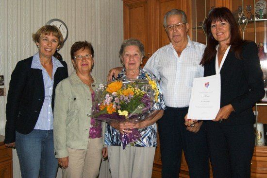 Fünf Menschen stehen in einem Raum zusammen. Die Frau in der Mitte hält einen Blumenstrauß. Die Frau auf der rechten Seite hält ein Zertifikat. Der Mann auf der linken Seite trägt eine Brille. Die Frau auf der linken Seite trägt eine Halskette. Die Wand hinter ihnen hat eine Uhr.