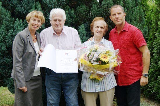Vier Personen stehen vor einer Hecke und lächeln. Der Mann in der Mitte hält ein Zertifikat, und die Frau neben ihm hält einen Blumenstrauß. Die Frau links trägt einen grauen Anzug, und der Mann rechts trägt ein rotes Hemd.
