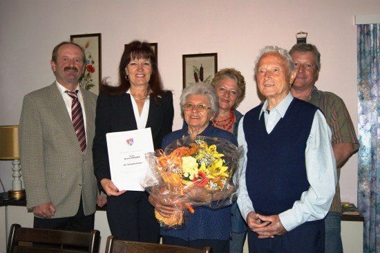 Eine Gruppe von Menschen steht in einem Raum zusammen, wobei eine Frau einen Blumenstrauß und ein Zertifikat hält. Die Frau lächelt und hält den Strauß mit beiden Händen. Hinter ihr steht ein Mann mit gefalteten Händen vor sich. Links steht ein Mann mit einer Krawatte und einer Lampe auf dem Tisch.