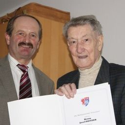 Zwei Männer stehen zusammen, einer hält ein Zertifikat mit einem Logo, beide lächeln für ein Foto.
