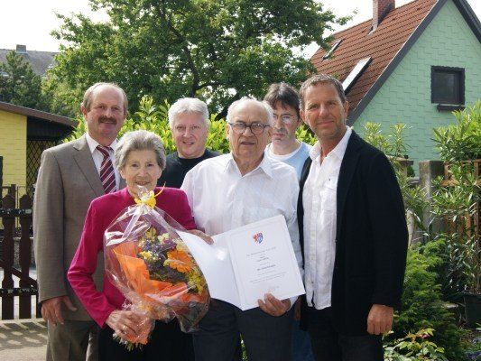 Eine Gruppe von Menschen steht vor einem Haus, ein Mann hält einen Blumenstrauß und ein Zertifikat, während ein anderer Mann ein Papier in der Hand hat.