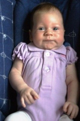 Ein Babymädchen mit hellbraunen Haaren trägt ein lila Kleid und sitzt auf einem blauen Kissen. Sie schaut mit leicht geöffnetem Mund in die Kamera.