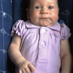 Ein Babymädchen mit hellbraunen Haaren trägt ein lila Kleid und sitzt auf einem blauen Kissen. Sie schaut mit leicht geöffnetem Mund in die Kamera.