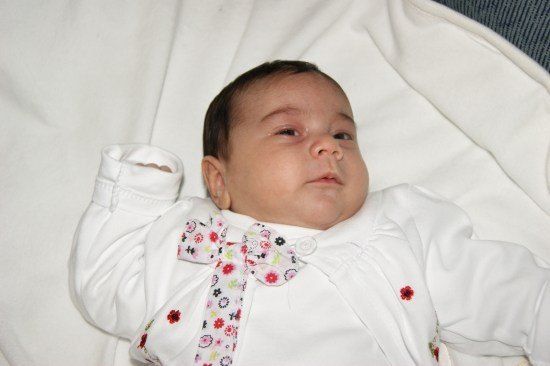 Ein Baby liegt auf einer weißen Decke und trägt ein weißes Outfit mit einer Blumenschleife und roten Blumen auf der Vorderseite. Die Augen des Babys sind leicht geschlossen, und es schaut nach links.