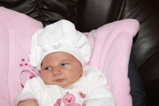 Ein Babymädchen in einer weißen Haube und einem rosa Outfit sitzt auf einem rosa Kissen auf einem schwarzen Ledersofa.