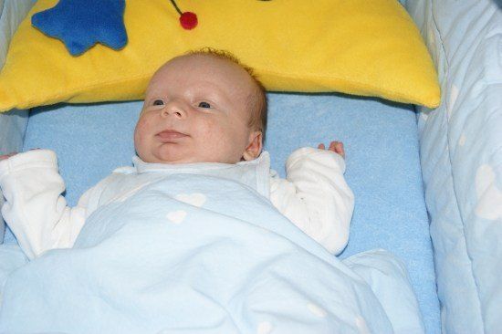 Ein Baby liegt in einem Kinderbett mit einer blauen Decke und einem gelben Kissen mit einem Stern und einer roten Kirsche obenauf.