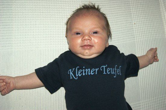 Ein Baby liegt auf einer weißen Oberfläche und trägt ein schwarzes T-Shirt mit blauem Text, der 'Kleiner Teufel' lautet. Das Baby hat leicht geöffneten Mund und schaut direkt in die Kamera.