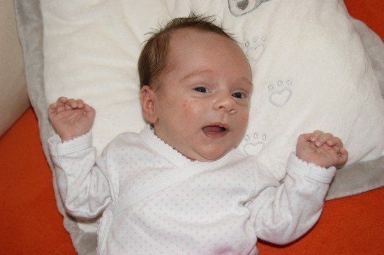 Ein Baby liegt auf einer Decke mit herzförmigem Design, schaut nach oben und lächelt mit beiden Händen erhoben.