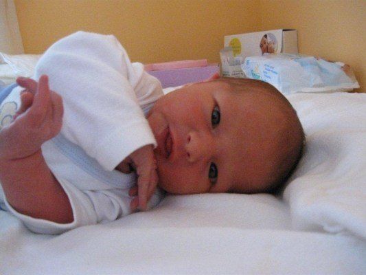 Ein Baby liegt auf einem Bett, in Weiß gekleidet, mit einem leichten Lächeln, schaut in die Kamera.