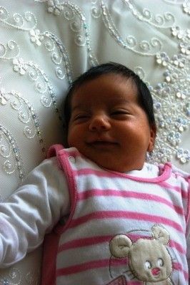 Ein Babymädchen liegt auf einem Bett mit einem weiß-rosa gestreiften Lätzchen, lächelt und schaut nach oben. Sie hat kurzes schwarzes Haar und trägt ein rosa-weißes Outfit mit einem Bärendesign.