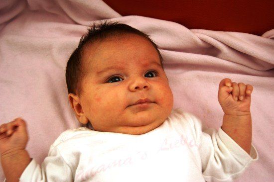 Ein Baby liegt auf einer rosa Decke, trägt ein weißes T-Shirt mit pinker Schrift und schaut direkt in die Kamera mit einem neugierigen Ausdruck.