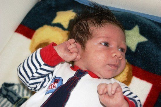 Ein Baby liegt auf einer Decke mit Sternmuster, trägt ein weiß-rot gestreiftes Shirt und eine Krawatte. Das Haar des Babys ist etwas zerzaust, und es hat einen ernsten Ausdruck.