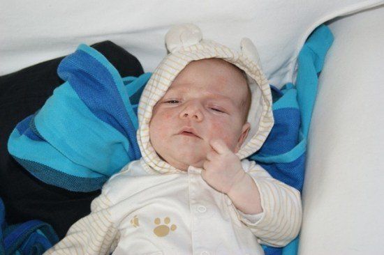 Ein Baby liegt auf einem Bett, eingewickelt in eine blaue Decke, und trägt eine weiße Kapuzenjacke mit einem Bärendesign auf der Vorderseite. Die Augen des Babys sind geschlossen, und es hat eine Hand am Mund.